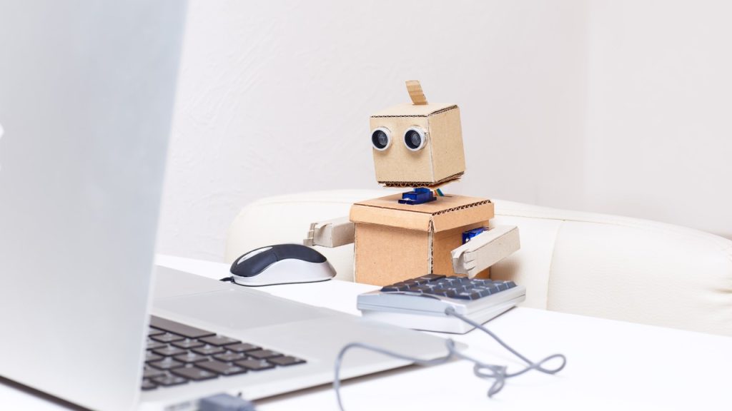 A small cardboard robot sat at a desk using a laptop, mouse and calculator.