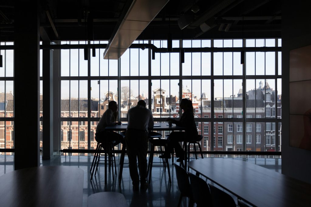 Three people sat at a table on high stools, silhouetted against a view through windows overlooking the top of a city street.