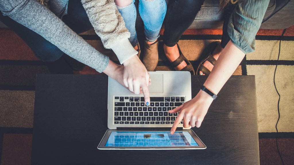 Three people sat on a sofa pointing at the keyboard and screen of an Apple Mac - viewed from above.